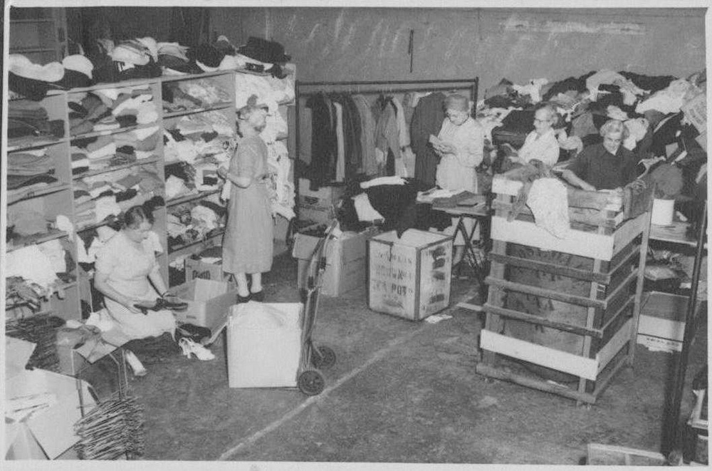 This is a black and white photo of a few female volunteers sorting clothes and other second-hand items in a warehouse facility.